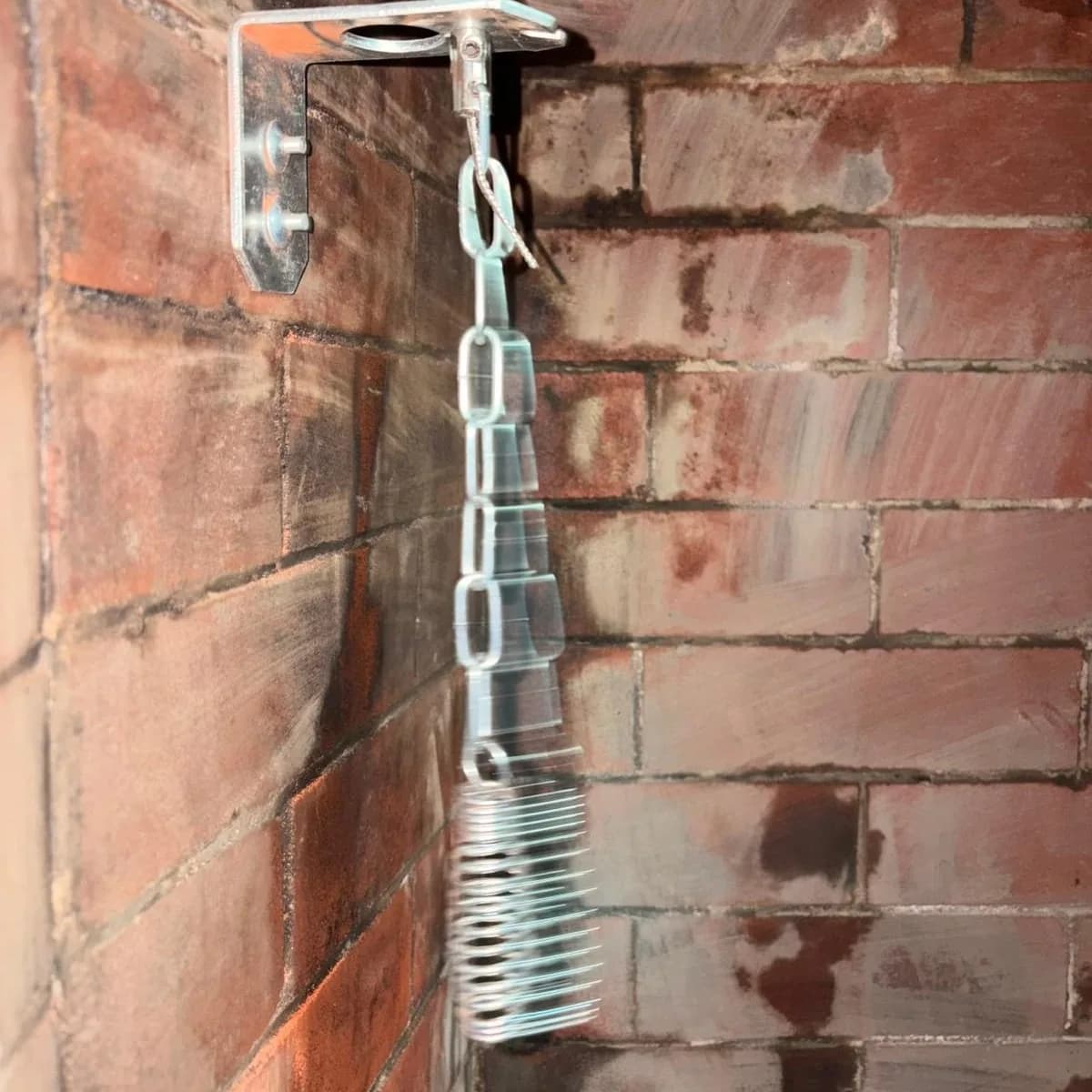A galvanised bracket bolted into firebrick inside a firebox, carrying a chain and coil spring for a top-sealing damper